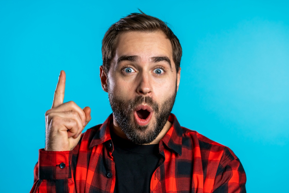 Portrait of young thinking pondering man with beard having idea moment pointing finger up on blue studio background. Smiling happy guy showing eureka gesture. Portrait of thinking pondering man beard having idea moment pointing finger up