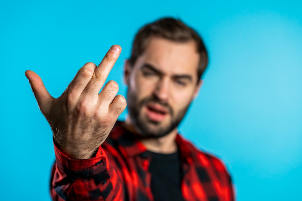 Young handsome man on blue background showing middle finger - gesture of fuck. Expression negative, aggression, provocation. Young handsome man on blue background showing middle finger - gesture of fuck. Expression negative, aggression, provocation.