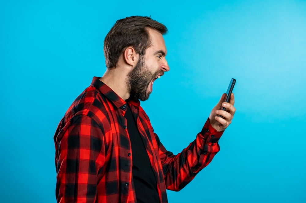 Angry modern hipster in red plaid shirt screaming down his mobile phone. Stressed and depressed man on blue background.. Angry modern hipster in red plaid shirt screaming down his mobile phone. Stressed and depressed man on blue background