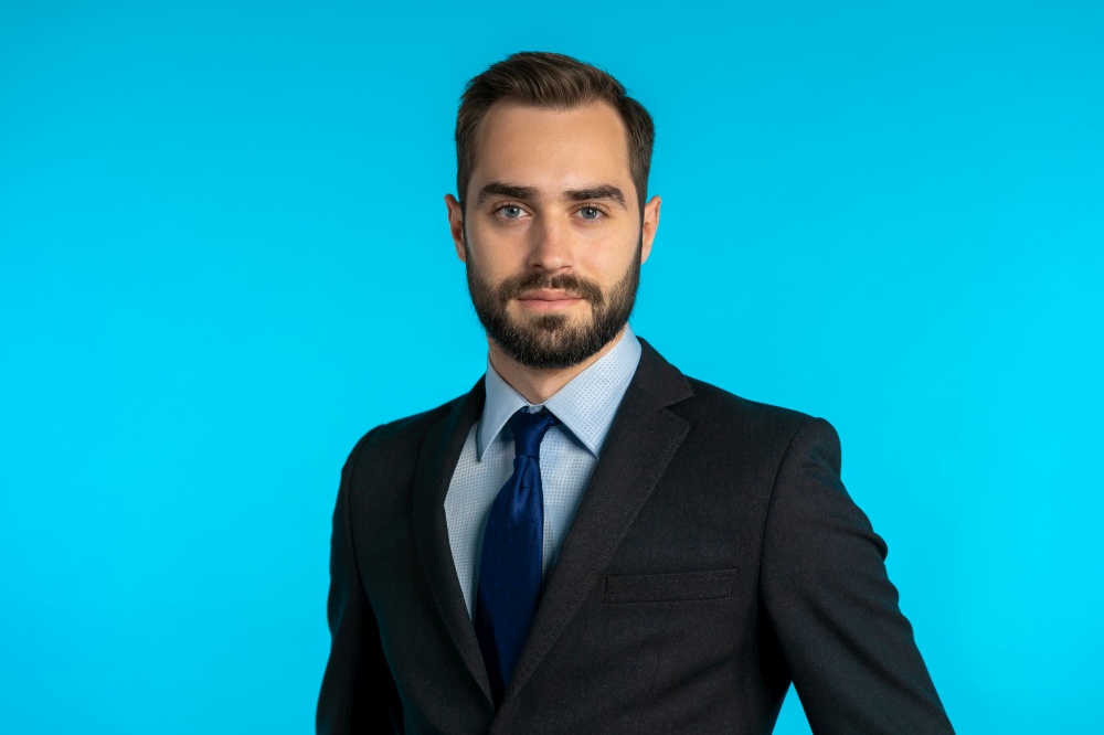 Close up portrait of young successful confident businessman with beard isolated on blue studio background. Man in business suit looking to camera and smiling. Portraiture of handsome guy. portrait of young successful confident businessman with beard isolated on blue
