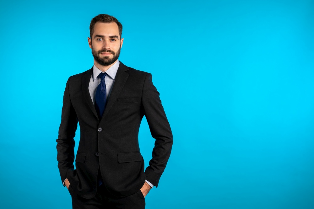 Close up portrait of young successful confident businessman with beard isolated on blue studio background. Man in business suit looking to camera and smiling. Portraiture of handsome guy. Copy space. Portrait of young successful confident businessman with beard isolated on blue studio background. Man in business suit looking to camera and smiling. Portraiture of handsome guy.