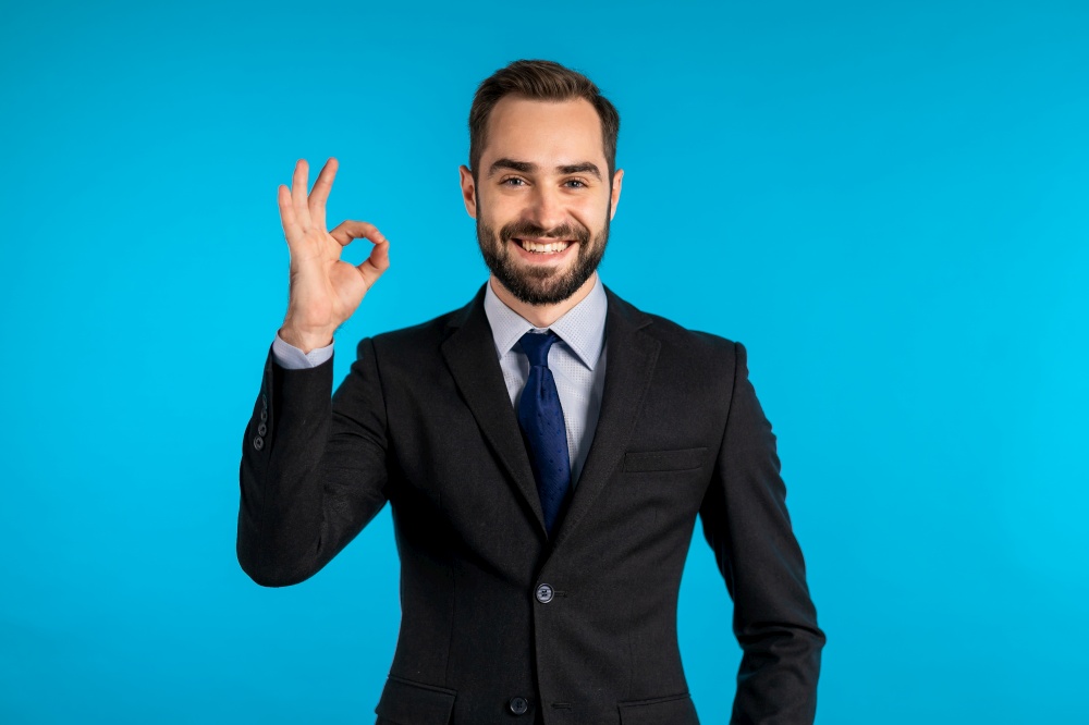 Positive young businessman smiles to camera. Man showing OK sign over blue background. Winner. Success. Body language. Positive young businessman smiles to camera. Man showing OK sign over blue background. Winner. Success. Body language.