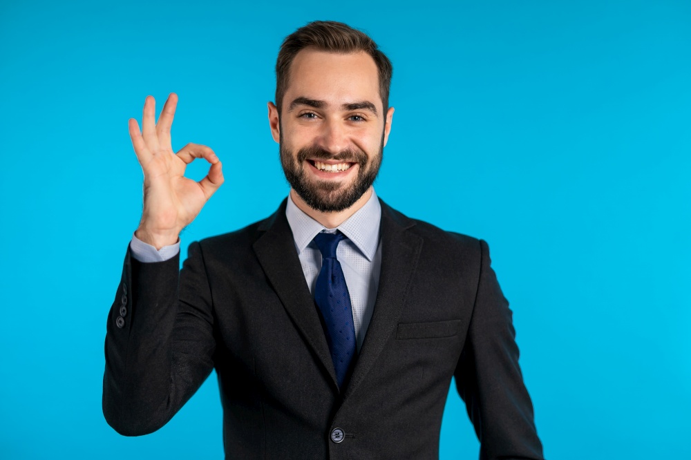 Positive young businessman smiles to camera. Man showing OK sign over blue background. Winner. Success. Body language. Positive young businessman smiles to camera. Man showing OK sign over blue