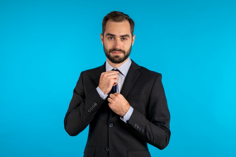 Handsome young businessman corrects his necktie and looking to camera like in mirror. Man on blue studio background. Corporate person in elegant suit correcting his tie. Handsome young businessman corrects his necktie and looking to camera