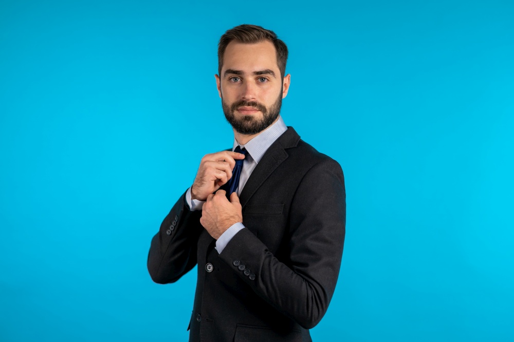 Handsome young businessman corrects his necktie and looking to camera like in mirror. Man on blue studio background. Corporate person in elegant suit correcting his tie. Handsome young businessman corrects his necktie and looking to camera like in mirror. Man on blue studio background. Corporate person in elegant suit correcting his tie.
