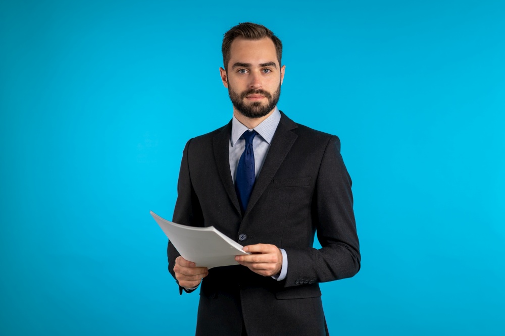 Young handsome man in suit jacket with documents, bills, report or contract. Businessman isolated on blue background. He is satisfied. Young handsome man in suit jacket with documents, bills, report or contract. Businessman isolated on blue background. He is satisfied.