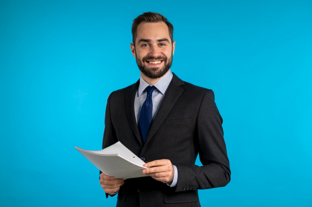 Young handsome man in suit jacket with documents, bills, report or contract. Businessman isolated on blue background. He is satisfied. Young handsome man in suit jacket with documents, bills, report or contract. Businessman isolated on blue background. He is satisfied.