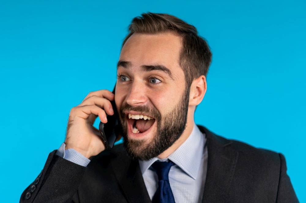 Surprised excited happy businessman talking on mobile telephone on blue background. Man saying wow. Surprised excited happy businessman talking on mobile telephone on blue