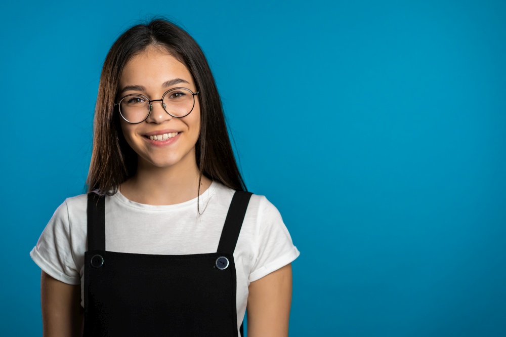 Pretty young asian girl with long hair standing on blue studio background. Cute portrait of woman in transparent round glasses and black overalls. Pretty young asian girl with long hair standing on blue studio background. Cute portrait of woman in transparent round glasses and black overalls.