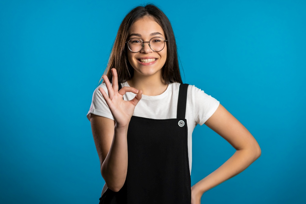 Young asian woman with perfect make-up making OK sign over blue background. Winner. Success. Positive girl smiles to camera. Body language. Young asian woman with perfect make-up making OK sign over blue background. Winner. Success. Positive girl smiles to camera. Body language.