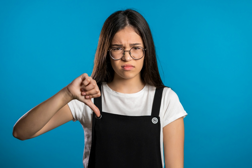 Young asian girl standing on blue studio background expressing discontent and showing thumb down gesture at camera. Portrait of woman with sign of dislike. Young asian girl standing on blue studio background expressing discontent and showing thumb down gesture at camera. Portrait of woman with sign of dislike.
