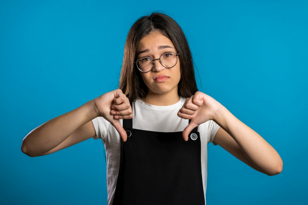 Young asian girl standing on blue studio background expressing discontent and showing thumb down gesture at camera. Portrait of woman with sign of dislike. Young asian girl standing on blue studio background expressing discontent and showing thumb down gesture at camera. Portrait of woman with sign of dislike.