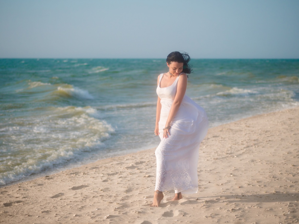 The young girl enjoys good windy weather on sea beach. Woman is dressed in white sundress and boho jewelry rest at the sea. Carefree and freedom concept. Portrait of gypsy lady.