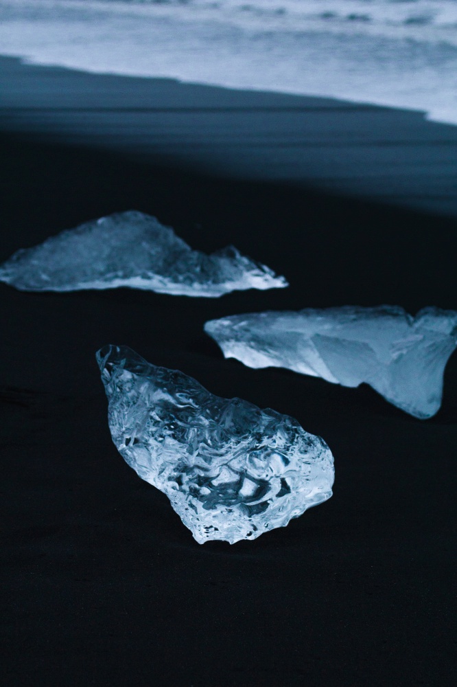 Close up transparent ice pieces on dark sand concept photo. Ocean beach. Front view photography with sea waves on background. High quality picture for wallpaper, travel blog, magazine, article. Close up transparent ice pieces on dark sand concept photo