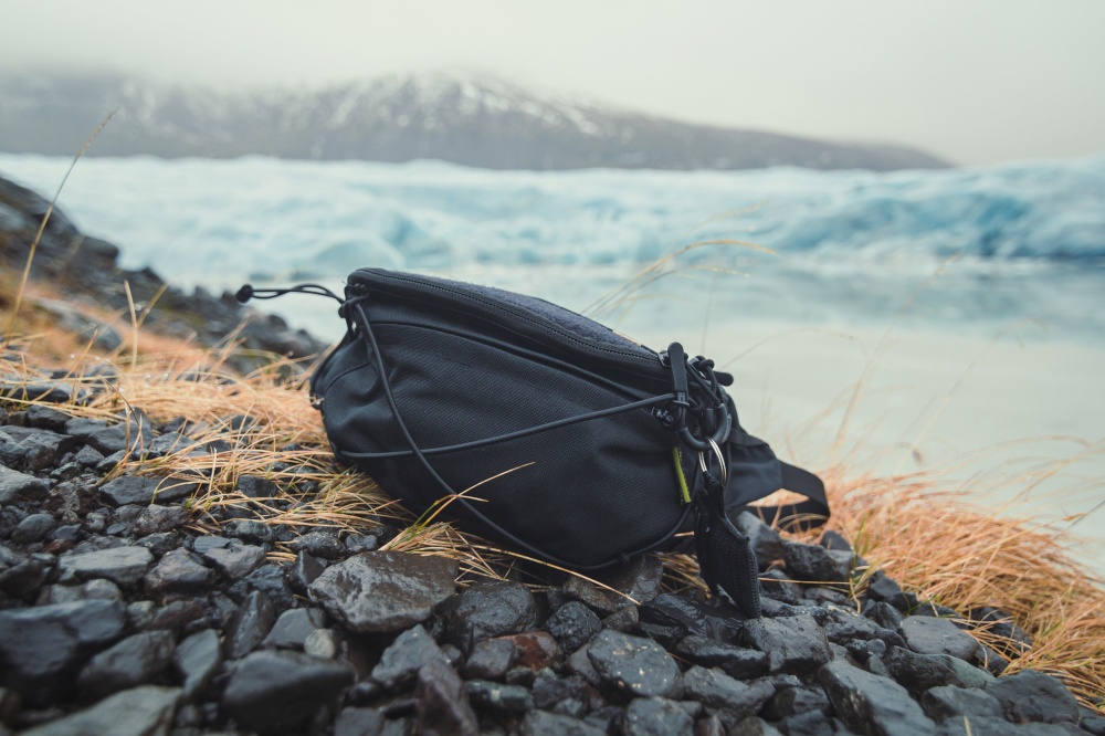 Close up camera bag on stones concept photo. Cameraman accessory outdoor. Front view photography with ice mountains on background. High quality picture for wallpaper, travel blog, magazine, article. Close up camera bag on stones concept photo