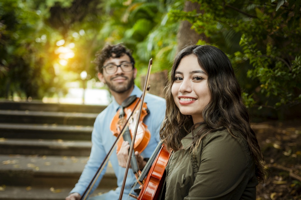 Two young violinists sitting outdoors looking at the camera. Portrait of two young violists sitting outside. Man and woman violinist sitting on the stairs. Concept of two young violinists