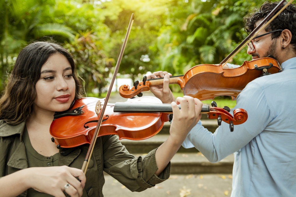 Violinist man and woman back to back playing violin in a park outdoors. Two young violinists standing playing violin in a park. Portrait of man and woman together playing violin in park