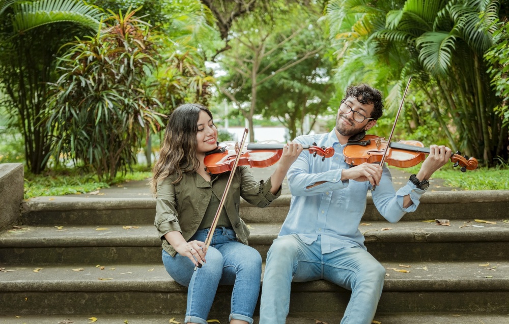 Portrait of two young violists playing violin sitting on stairs outside. Portrait of smiling violinists sitting on stairs playing violin. Man and woman sitting on stairs playing violin