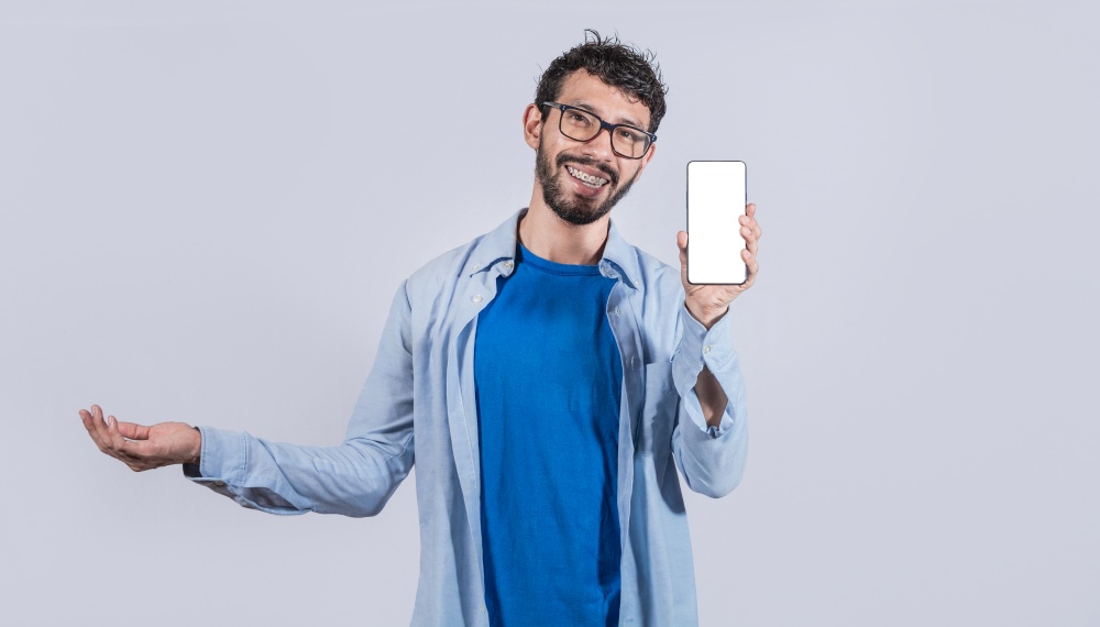 Handsome man smiling positive showing mobile phone blank screen with another hand palm open, excited happy male holding smartphone