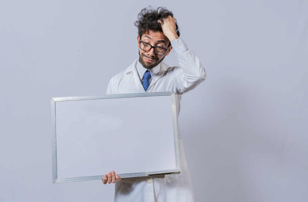 Male scientist in white coat holding and pointing at a blank whiteboard. Disheveled-haired man holding blank whiteboard, Amazed man in white coat pointing at a whiteboard