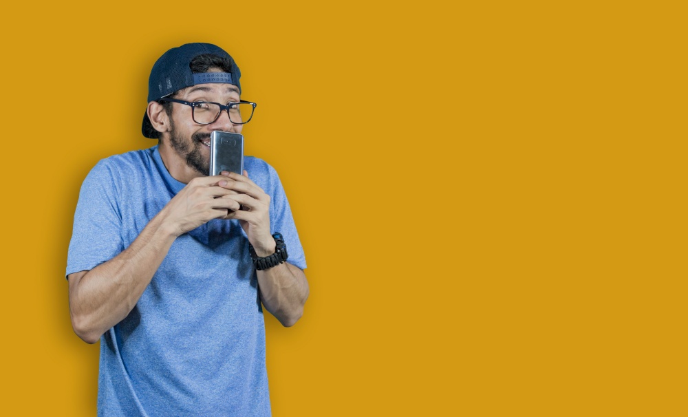 boy in glasses smiling with his cell phone, young man in cap smiling while holding his cell phone on yellow background, boy smiling covering his mouth with cell phone