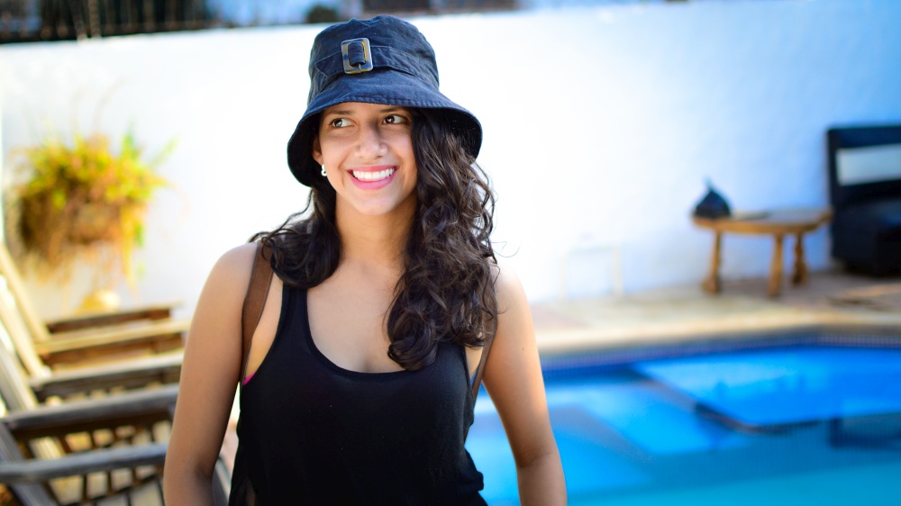 Pretty girl with hat smiling near a swimming pool, latin girl smiling by a swimming pool with muskoka chairs