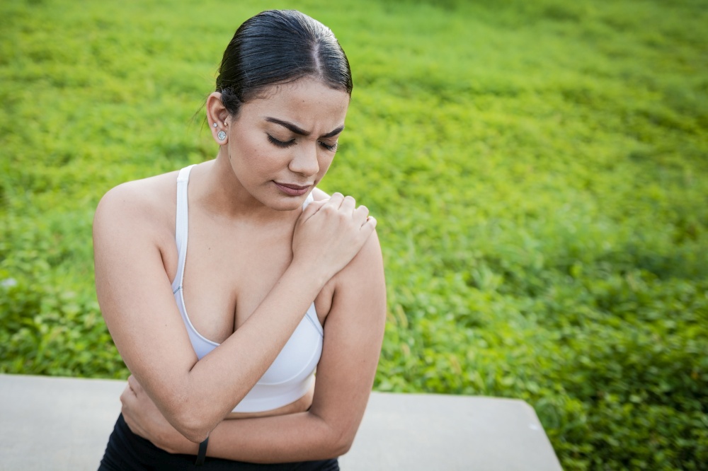 Girl suffering from shoulder pain sitting outdoors. Woman sitting and stressed with shoulder pain outdoors. Girl with shoulder pain outdoors, Young woman with shoulder pain sitting outside