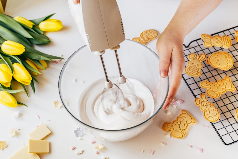 Cropped shot of woman using mixer machine making cookie icing for decoration. Woman holding glass bowl with sugar glaze and electric mixer. Easter holiday backing. Woman whisk cookie icing for decoration