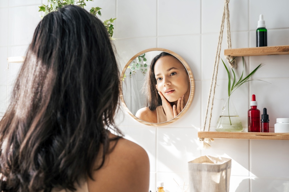 Beautiful woman with dark skin looking in the mirror in white eco friendly bathroom. Wooden shleves and reusable cosmetics bottles. Wellnes concept. Beautiful woman looking in the mirror