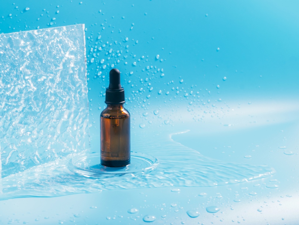A bottle with a pipette, a cosmetic product, splashes and drops of water on a blue background. Face, body, hair care, moisturizing nutrition. Horizontal frame copy space, branding. A bottle with a pipette, a cosmetic product, splashes and drops of water on a blue background