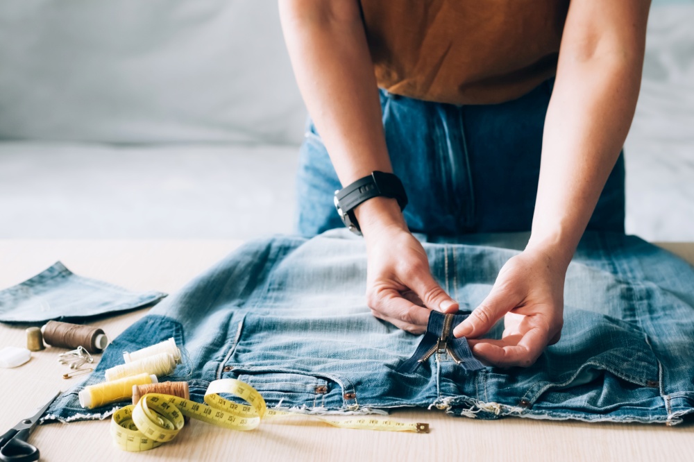 Woman repairs sews reuses fabric from old denim clothes. The concept of economical reuse of recyclable things. Homemade needlework hobby. Selective focus. Woman repairs sews reuses fabric from old denim clothes economical reuse