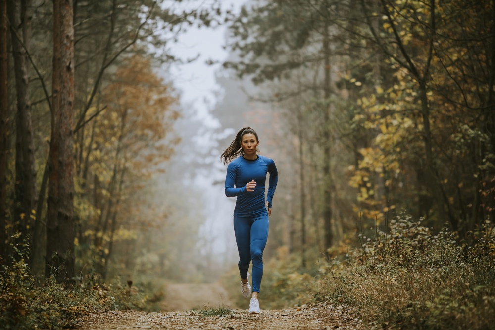 Young woman in blue track suit running toward camera on the forest trail at autumn