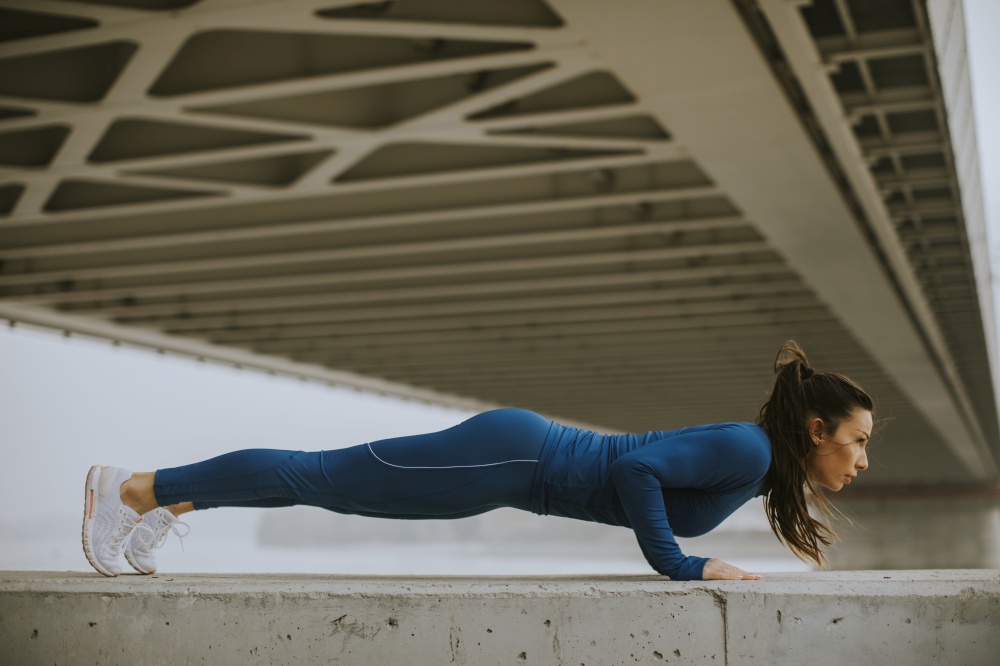 Young woman in blue track suit doing pushups under the bridge in the urban environment
