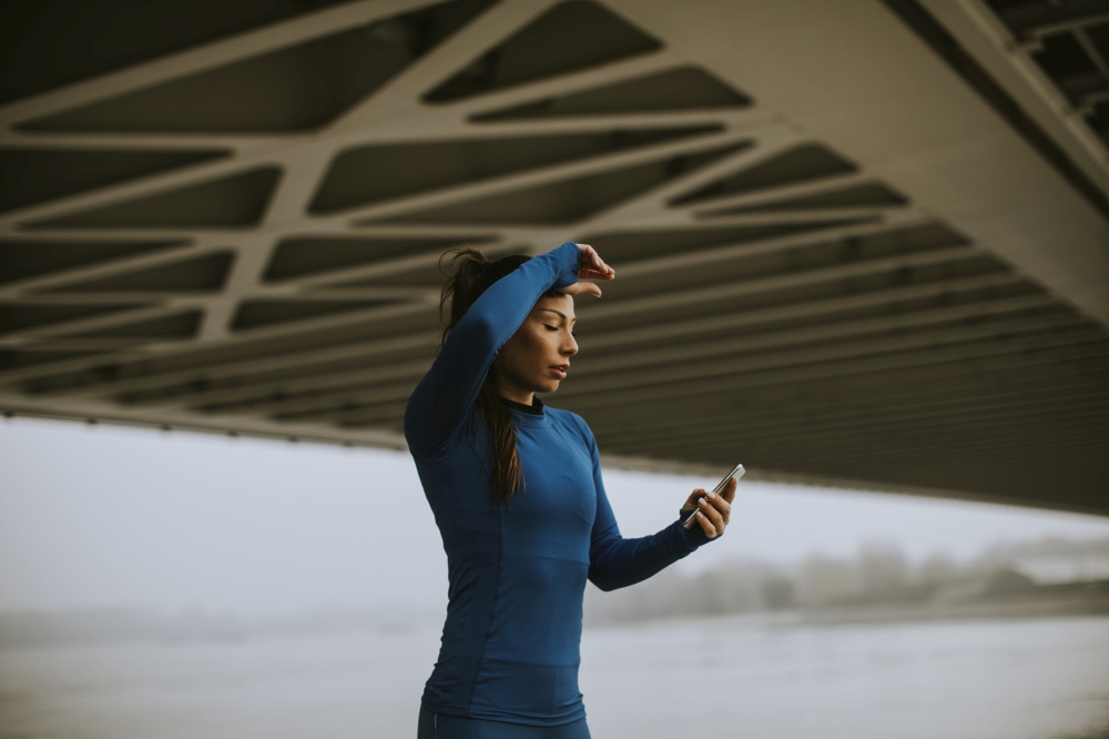 Pretty young woman in blue track suit monitoring her workout progress on fitness app by the river at autumn morning