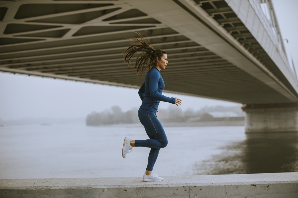 Pretty young woman in blue track suit running by the river at autumn morning
