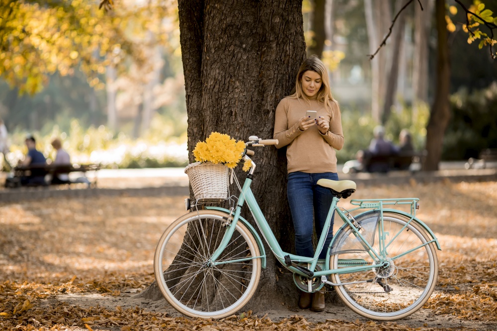 Pretty young woman with bicycle using smartphone in autumn park