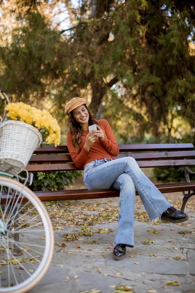 Pretty young woman using phone in autumn park sitting on bench by the bicycle