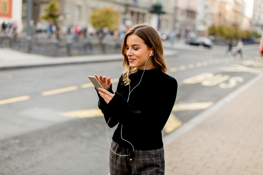 Smiling brunette woman using mobile while standing on street and  waiting for a bus or taxi