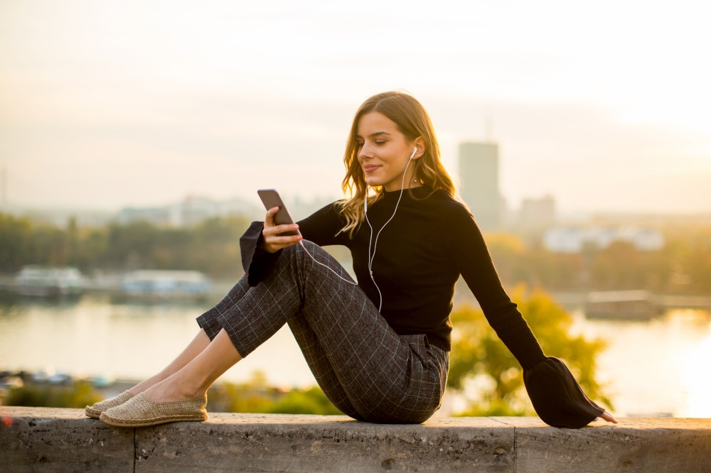 Portrait of trendy young woman listening music from smartphone outdoor at sunset