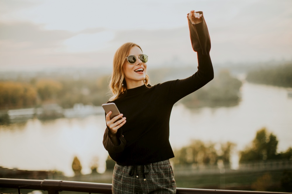 Portrait of young woman holding mobile phone while standing on walkway by the river