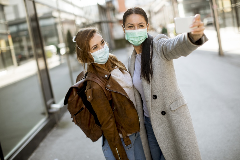 Two pretty young women with protective facial mask using mobile phone to take sephie on the street