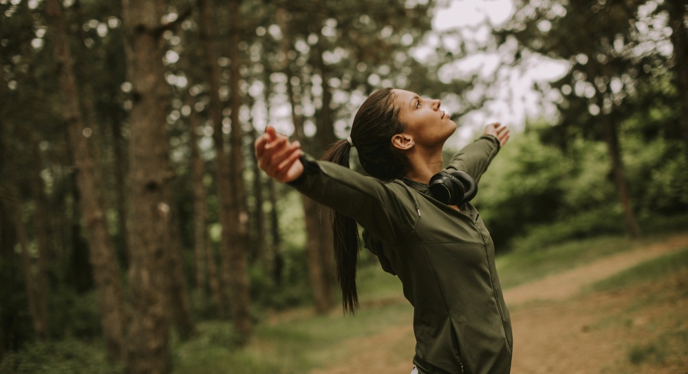 Pretty young woman with earphones preading her arms in the forest because she enjoys training outside