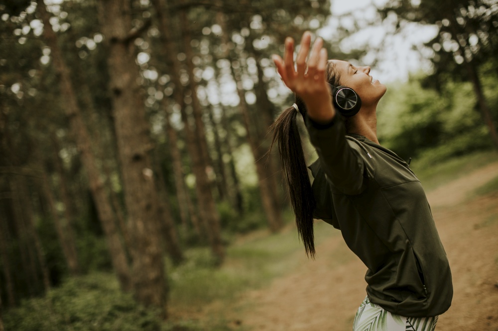 Pretty young woman with earphones preading her arms in the forest because she enjoys training outside