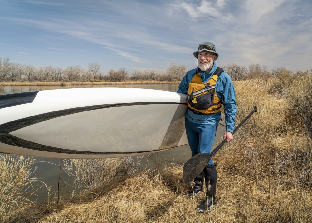 happy senior male paddler with his paddleboard and paddle on a lake shore in Colorado in early spring scenery