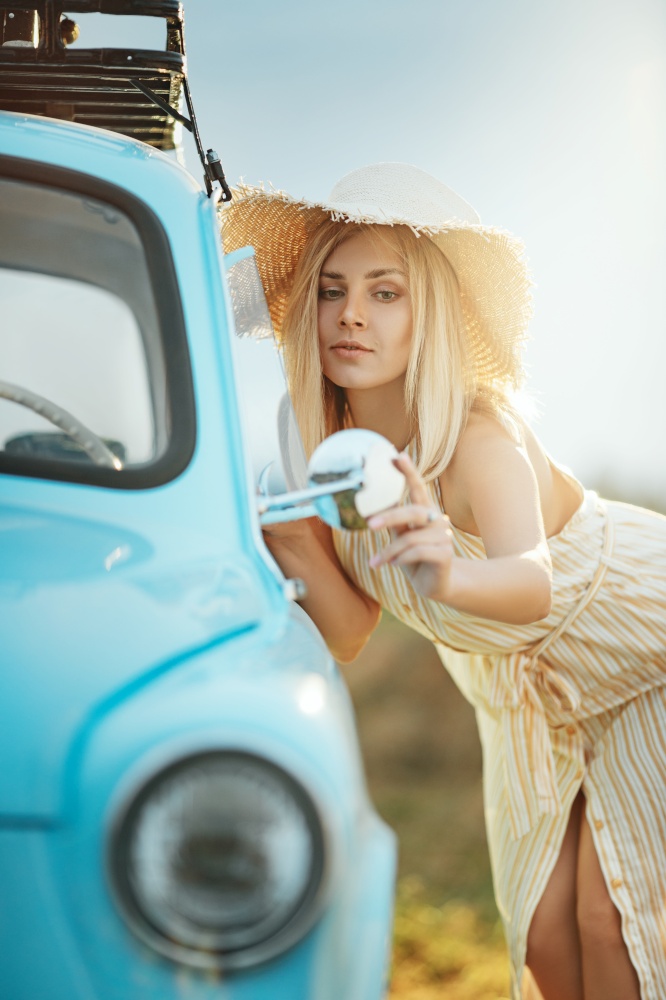 Young woman in straw hat standing near a vintage car and looking at rear view mirror, summer vibe