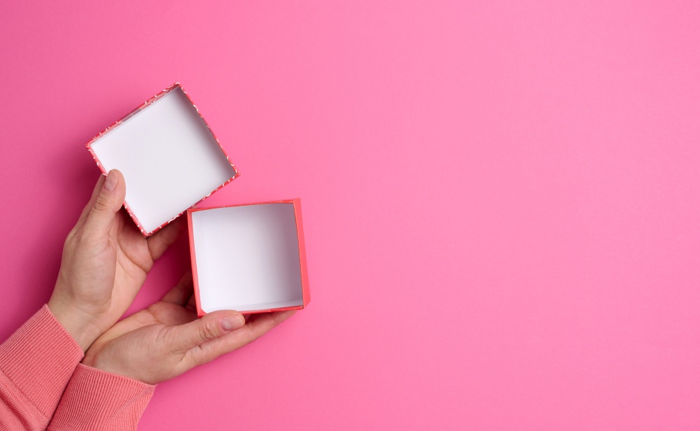 female hands holding open square cardboard gift box on pink background, top view