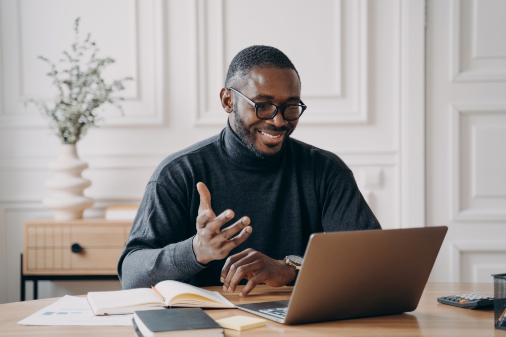 African-American young businessman in glasses having conference online with employes via video call broadly smiling while looking at laptop screen gesturing when communicates and answering questions. African-American young businessman in glasses having conference online with employes via video call