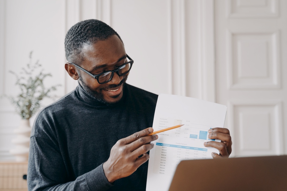 Young smiling african american businessman in glasses holding document with graphs, explaining financial report during online meeting with colleagues on laptop, sitting at desk at home office. Young smiling african american businessman explaining financial report during online meeting