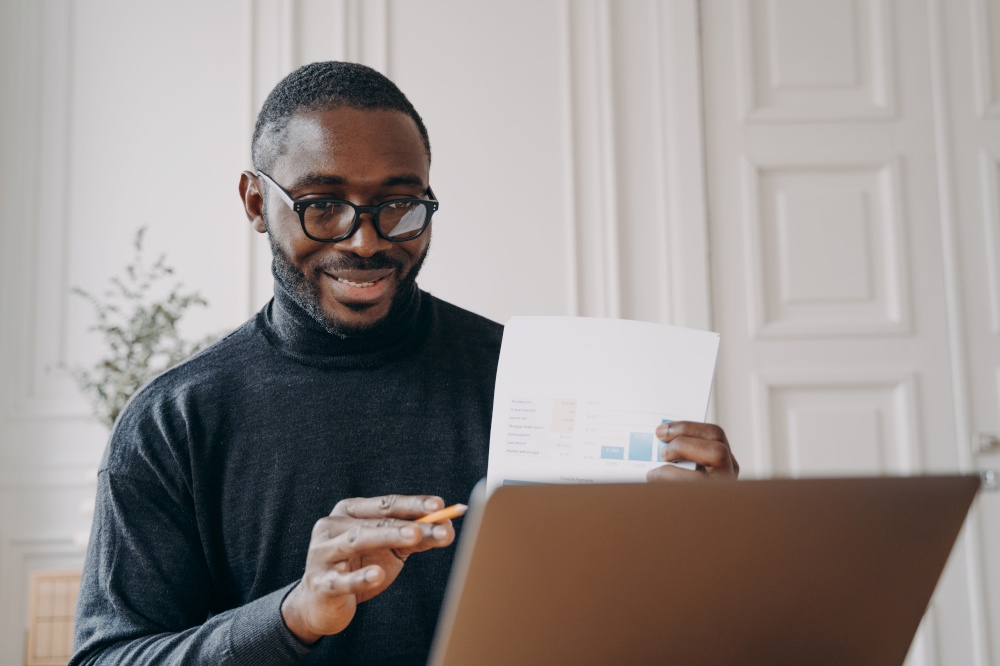 Positive young african american man freelancer in glasses having video conference with partner on laptop while working remotely at home, holding financial report in hand during online meeting. Positive young african american man freelancer having video conference with partner on laptop