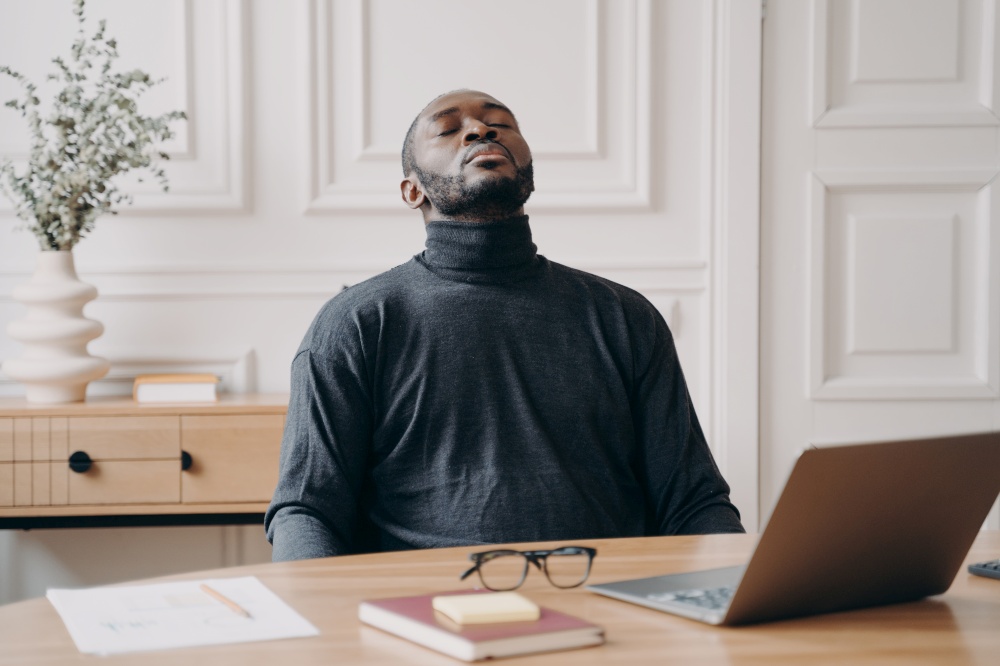 Exhausted home office employee Afro American male sitting in front of laptop taking off glasses closing eyes and throwing head back. Tired young man trying to recover to continue remote work online. Exhausted home office employee African american male sitting with eyes closed throwing his head back
